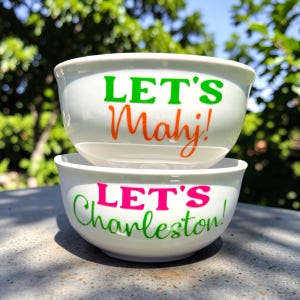 May include: Two white ceramic bowls stacked on top of each other. The top bowl has the text "LET'S Mahj!" in green and orange. The bottom bowl has the text "LET'S Charleston!" in pink and green. The bowls are outdoors.