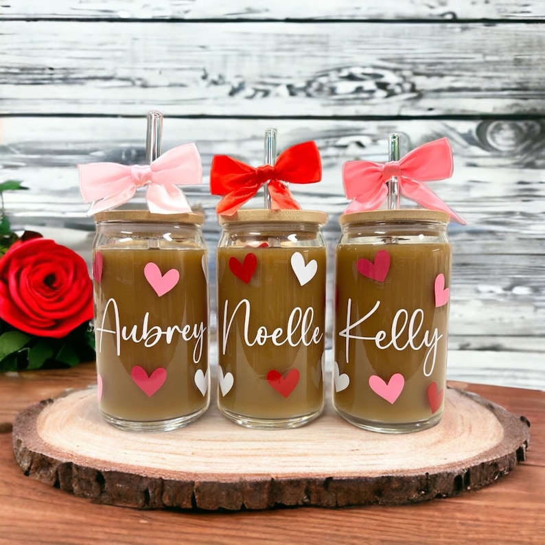May include: Three glass cups with bamboo lids and pink and red bows. Each cup has a brown liquid inside and is decorated with white and pink hearts. The cups are labeled "Aubrey", "Noelle", and "Kelly".
