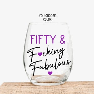May include: A clear stemless wine glass with the words "FIFTY & Fucking Fabulous" in purple and black script. The words "You Choose Color" are above the glass. The glass sits on a wooden surface.
