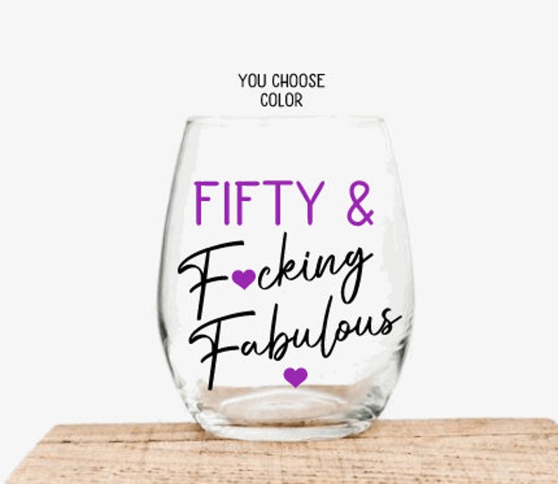 May include: A clear stemless wine glass with the words "FIFTY & Fucking Fabulous" in purple and black script. The words "You Choose Color" are above the glass. The glass sits on a wooden surface.