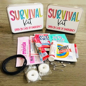 May include: Two white metal survival kit boxes with colourful text and contents including lip balm, wipes, and mints. The boxes read "SURVIVAL Kit" and "OPEN IN CASE OF EMERGENCY". The kit includes a black hair tie and safety pins.