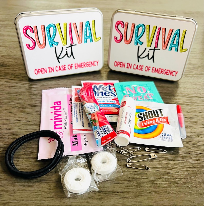 May include: Two white metal survival kit boxes with colourful text and contents including lip balm, wipes, and mints. The boxes read "SURVIVAL Kit" and "OPEN IN CASE OF EMERGENCY". The kit includes a black hair tie and safety pins.