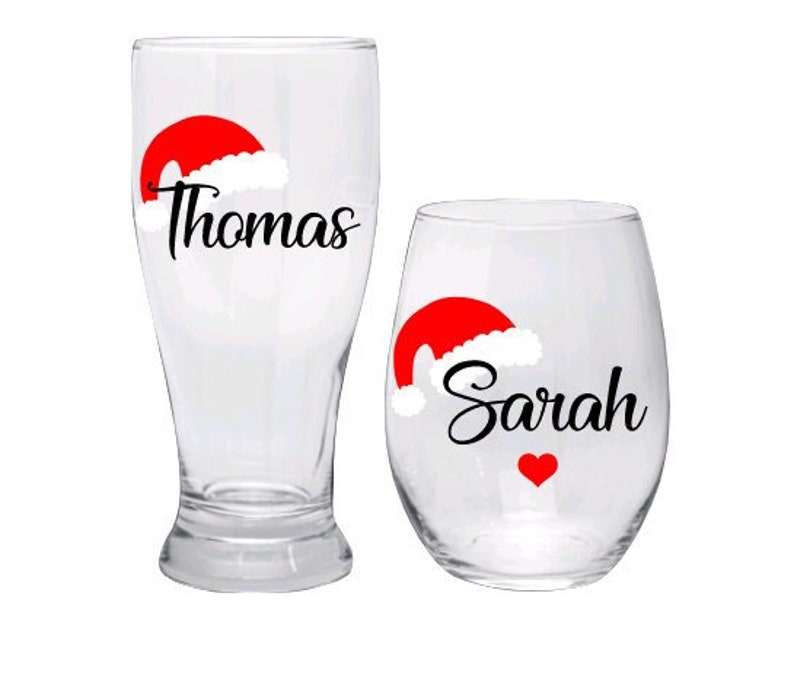 May include: Two clear glass beverage glasses with a red and white Santa hat design and personalized names "Thomas" and "Sarah".