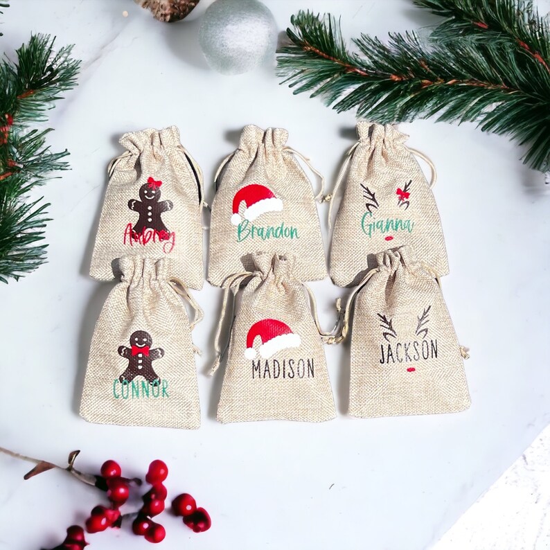 May include: Six small burlap gift bags with personalized names and Christmas-themed designs. The designs include a gingerbread person, Santa hat, and reindeer. The names on the bags are: Amy, Brandon, Gianna, Connor, Madison, and Jackson.