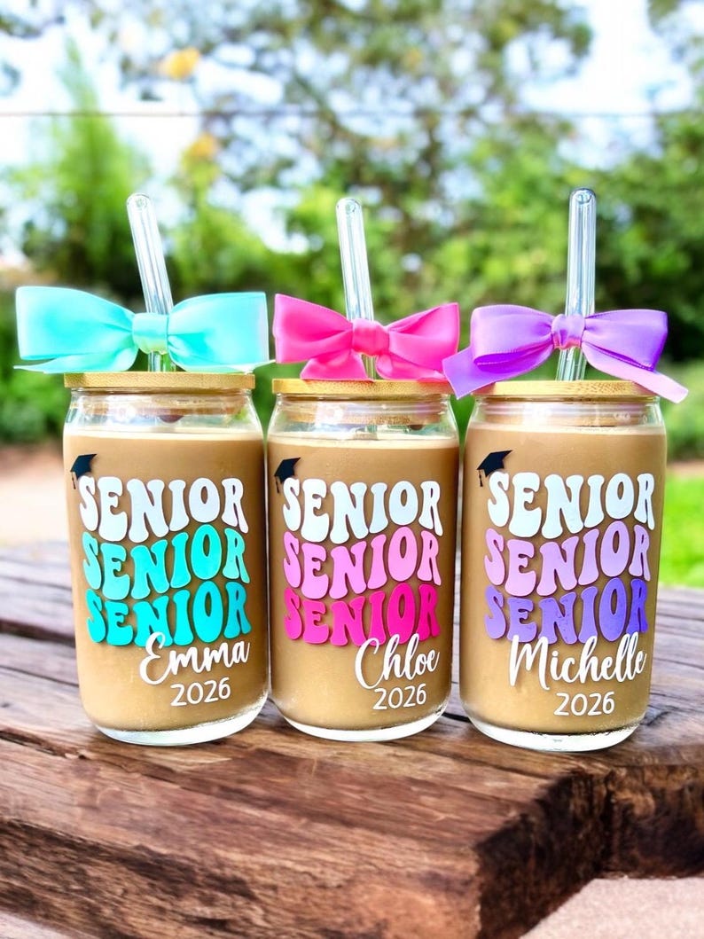 May include: Three glass tumblers with bamboo lids and clear glass straws, each topped with a colorful bow. The glasses feature the word SENIOR in stacked, wavy lettering, a small graduation cap icon, and the names Emma, Chloe, and Michelle with 2026.