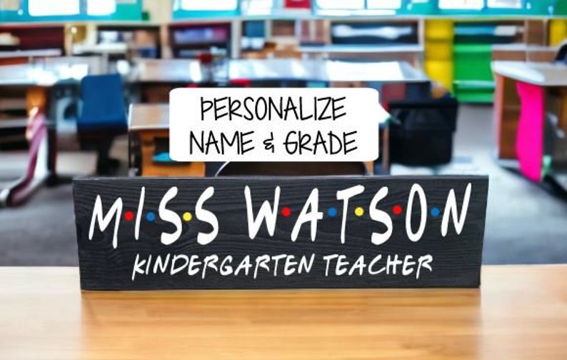 May include: Black chalkboard sign with white text that reads "MISS WATSON KINDERGARTEN TEACHER". The letters are in a stylized font with colorful dots between them. A white speech bubble above the sign reads "PERSONALIZE NAME & GRADE".