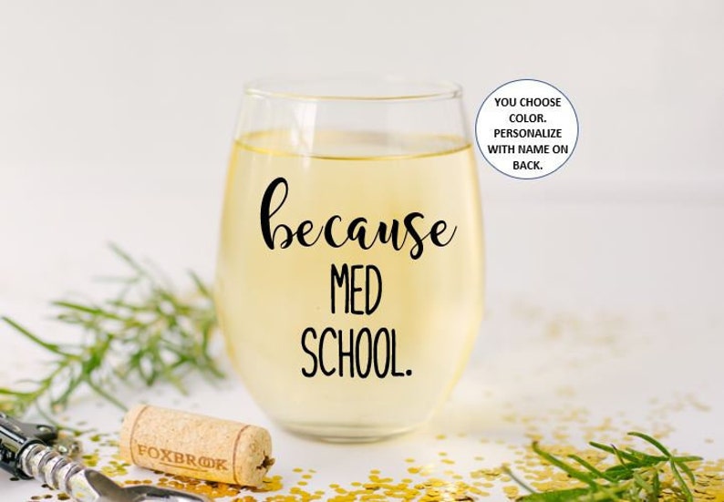 May include: A clear wine glass with the text "because med school." printed on it. The glass is filled with a light yellow liquid. The text is in black.