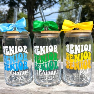 May include: Three clear glass tumblers with bamboo lids and glass straws. Each features a graduation cap icon, the word SENIOR repeated three times in a wavy font, a name, and the year 2026. They are adorned with blue, green, and yellow bows.