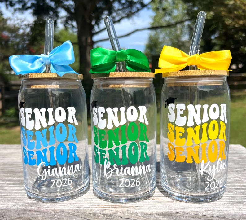 May include: Three clear glass tumblers with bamboo lids and glass straws. Each features a graduation cap icon, the word SENIOR repeated three times in a wavy font, a name, and the year 2026. They are adorned with blue, green, and yellow bows.