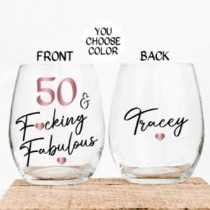 May include: Two clear stemless wine glasses with a rose gold design. The front glass says "50 & F*cking Fabulous" and the back glass says "Tracey".