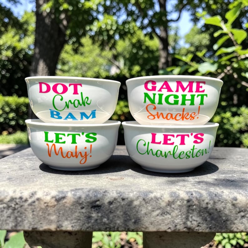 May include: Four white ceramic bowls stacked on a stone surface. Each bowl has colorful text: "DOT Crak BAM", "GAME NIGHT Snacks!", "LET'S Mahj!", and "LET'S Charleston!". The bowls are outdoors with a blurred green background.
