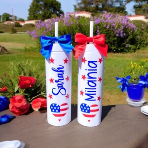 May include: Two white tumblers with red, white, and blue American flag heart designs and red stars. The tumblers have blue and red bows and straws. The tumblers are personalized with the names "Sarah" and "Milania".