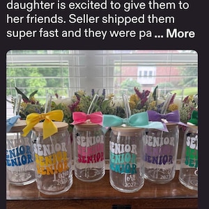 May include: A set of clear glass tumblers with bamboo lids and glass straws. Each tumbler features the word SENIOR printed in a wavy font in various colors, including yellow, pink, teal, purple, and green, with a matching satin bow tied to the straw.