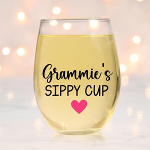 May include: A clear glass wine glass with a pink heart and the text "Gramie's Sippy Cup" in black lettering.