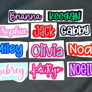 May include: A collection of custom name decals in various colors and fonts. Names include Brianna, Keegan, Sophia, Jack, Gabby, Miley, Olivia, Noah, Aubrey, Kaitlyn, and Noelle. Each name is on a white rectangular background.