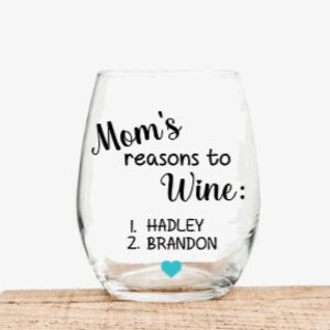 May include: A clear glass wine glass with a stem and a rounded base. The glass has a black inscription that reads "Mom's reasons to wine: 1. HADLEY 2. BRANDON" with a small blue heart.