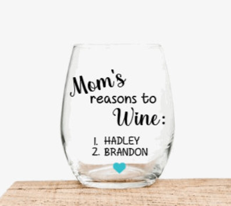 May include: A clear glass wine glass with a stem and a rounded base. The glass has a black inscription that reads "Mom's reasons to wine: 1. HADLEY 2. BRANDON" with a small blue heart.