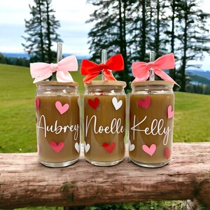 May include: Three glass cups with bamboo lids and pink, red, and white bows on the straws. The cups are filled with a brown liquid and have white hearts and pink hearts on them. The cups are labeled "Aubrey", "Noelle", and "Kelly".