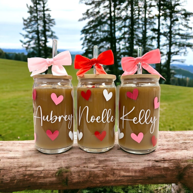 May include: Three glass cups with bamboo lids and pink, red, and white bows on the straws. The cups are filled with a brown liquid and have white hearts and pink hearts on them. The cups are labeled "Aubrey", "Noelle", and "Kelly".