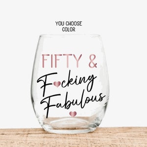 May include: A clear stemless wine glass with the words "FIFTY & Fucking Fabulous" in rose gold and black script. The text "YOU CHOOSE COLOR" is above the glass. The glass is sitting on a wooden surface.