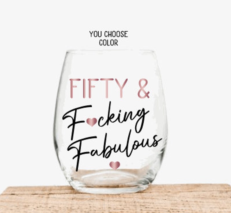 May include: A clear stemless wine glass with the words "FIFTY & Fucking Fabulous" in rose gold and black script. The text "YOU CHOOSE COLOR" is above the glass. The glass is sitting on a wooden surface.