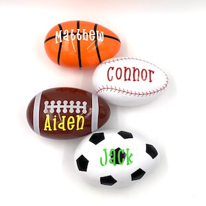 May include: Four white eggs decorated with sports themes. The eggs are decorated with a basketball, baseball, football, and soccer ball. Each egg has a name written on it: Matthew, Connor, Aiden, and Jack.