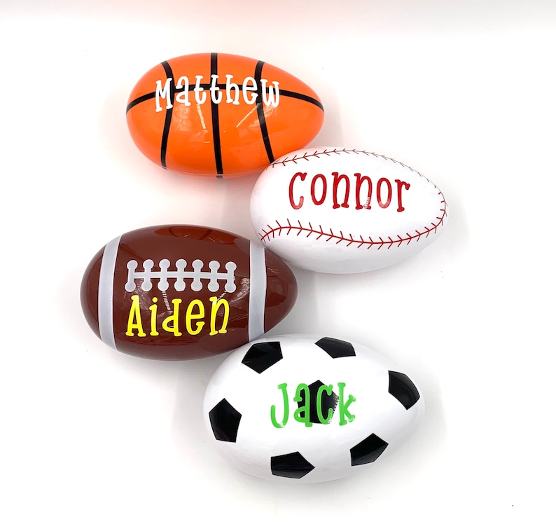May include: Four white eggs decorated with sports themes. The eggs are decorated with a basketball, baseball, football, and soccer ball. Each egg has a name written on it: Matthew, Connor, Aiden, and Jack.