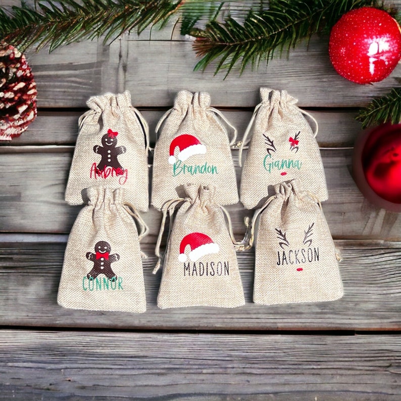 May include: Six small burlap gift bags with personalized Christmas designs. The bags feature a gingerbread man, Santa hat, and reindeer with names like "Anna", "Brandon", "Gianna", "Connor", "Madison", and "Jackson".