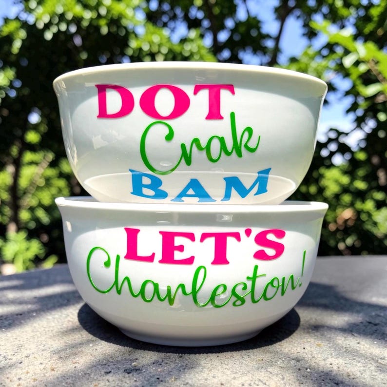 May include: Two white ceramic bowls stacked on top of each other. The top bowl has the words "DOT Crak BAM" in pink, green, and blue. The bottom bowl says "LET'S Charleston!" in pink and green.