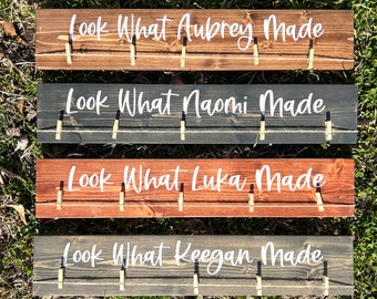 May include: Four wooden signs with the text "Look What [Name] Made" on each sign. The signs are painted in different colors, including brown, gray, and red. Each sign has a row of clothespins attached to the top.
