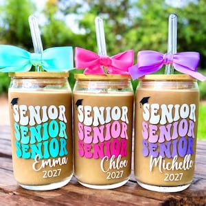 May include: Three glass tumblers with bamboo lids and clear glass straws, each topped with a satin bow in turquoise, pink, or purple. The glasses feature the text SENIOR in wavy letters, a graduation cap icon, and the names Emma, Chloe, or Michelle with 2027.