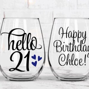 May include: Two clear stemless wine glasses. One glass has the text "hello 21" with blue heart accents. The other glass has the text "Happy Birthday Chloe!" in black script. Both glasses are on a white surface.
