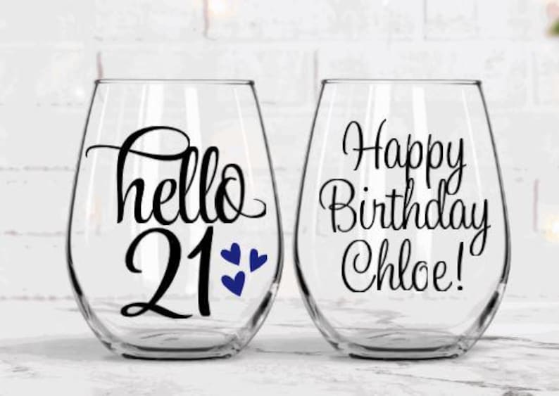 May include: Two clear stemless wine glasses. One glass has the text "hello 21" with blue heart accents. The other glass has the text "Happy Birthday Chloe!" in black script. Both glasses are on a white surface.