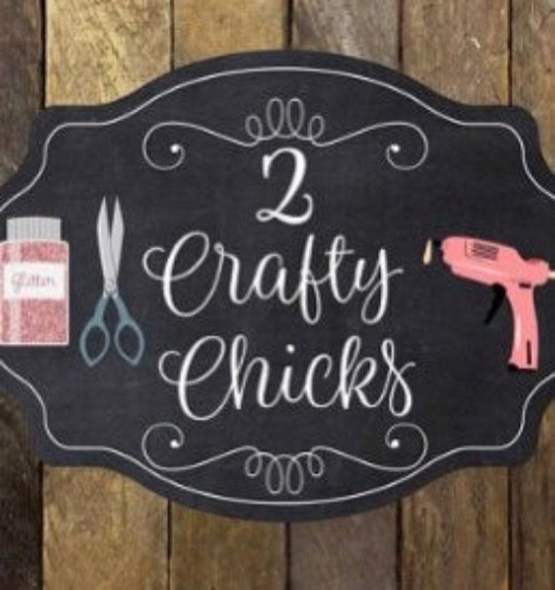 May include: Black chalkboard sign with white lettering that reads "2 Crafty Chicks". The sign has a decorative border and includes images of scissors, a glue gun, and a jar of glitter.
