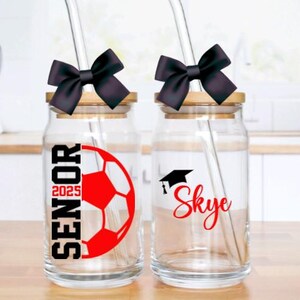 May include: Two glass jars with black lids and black bows. The jars have straws and are decorated with graduation caps, a soccer ball, and the text "Senior 2025" and "Skye".
