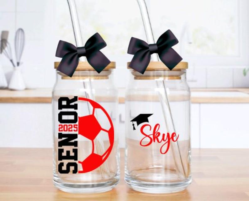 May include: Two glass jars with black lids and black bows. The jars have straws and are decorated with graduation caps, a soccer ball, and the text "Senior 2025" and "Skye".