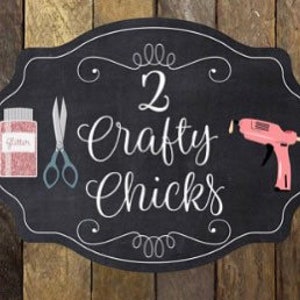 May include: A black chalkboard sign with a white border and the text "2 Crafty Chicks" in white cursive lettering. The sign is decorated with a pair of scissors, a jar of pink glitter, and a pink glue gun.