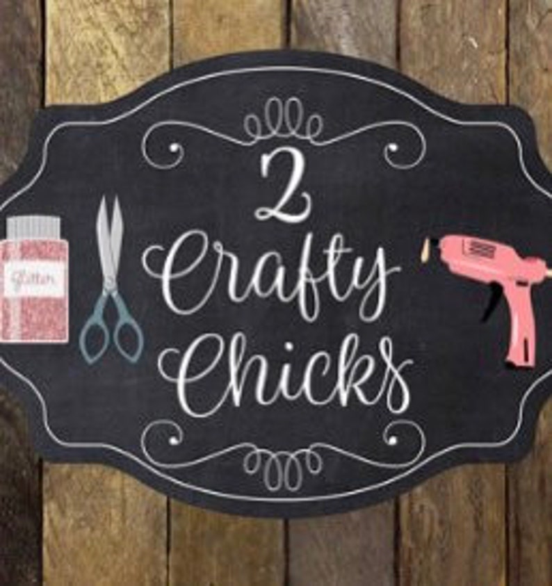 May include: A black chalkboard sign with a white border and the text "2 Crafty Chicks" in white cursive lettering. The sign is decorated with a pair of scissors, a jar of pink glitter, and a pink glue gun.