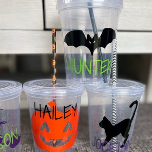 May include: Three clear plastic cups with black and white striped straws. The cups have Halloween-themed designs and names on them: "Hunter" with a black bat, "Hailey" with an orange pumpkin, and "Jocelyn" with a black cat.