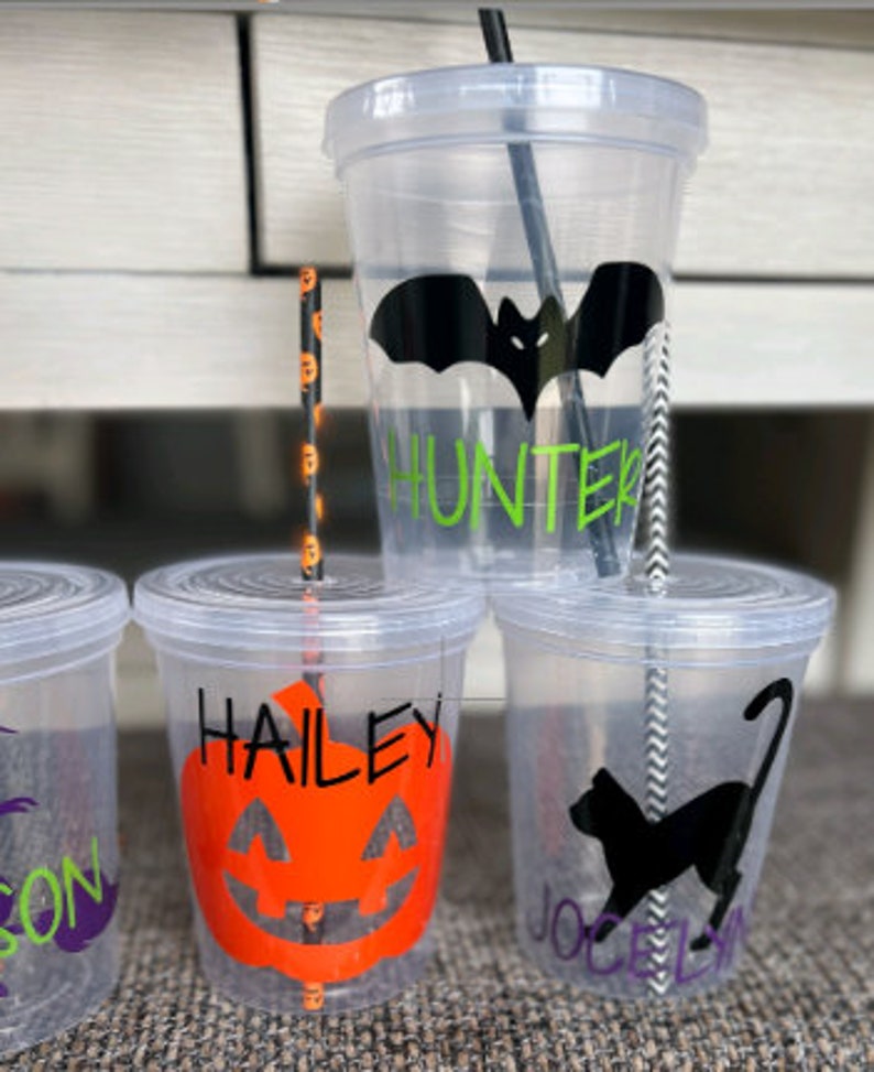 May include: Three clear plastic cups with black and white striped straws. The cups have Halloween-themed designs and names on them: "Hunter" with a black bat, "Hailey" with an orange pumpkin, and "Jocelyn" with a black cat.
