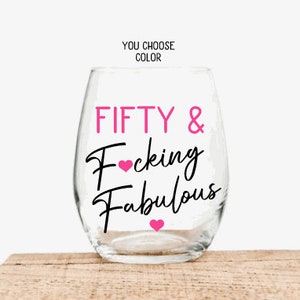 May include: A clear stemless wine glass with the text "FIFTY & Fucking Fabulous" in pink and black script. The words are separated by a black line and pink hearts. The words "YOU CHOOSE COLOR" are above the glass.