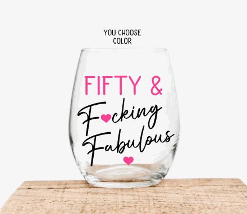 May include: A clear stemless wine glass with the text "FIFTY & Fucking Fabulous" in pink and black script. The words are separated by a black line and pink hearts. The words "YOU CHOOSE COLOR" are above the glass.