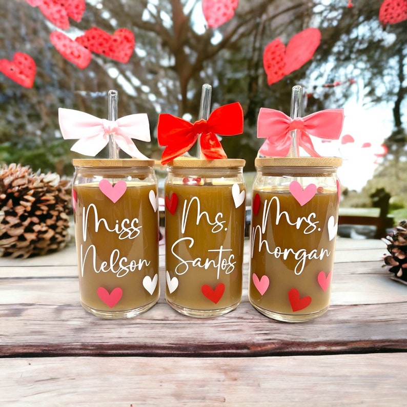 May include: Three glass cups with bamboo lids and straws. Each cup has a different name written on it: Miss Nelson, Mrs. Santos, and Mrs. Morgan. The cups are decorated with pink and red hearts.