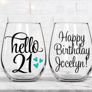May include: Two stemless wine glasses with black lettering. One glass says "hello 21" with teal hearts, the other says "Happy Birthday Jocelyn!". The glasses are clear and sit on a white surface.