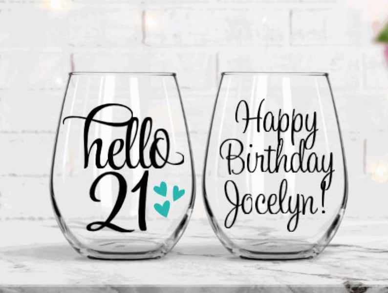 May include: Two stemless wine glasses with black lettering. One glass says "hello 21" with teal hearts, the other says "Happy Birthday Jocelyn!". The glasses are clear and sit on a white surface.