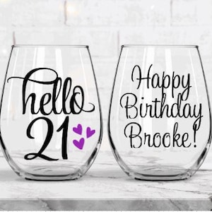 May include: Two clear stemless wine glasses. One glass has the words "hello 21" with purple heart accents. The other glass says "Happy Birthday Brooke!" in black script. The glasses are on a white marble surface.