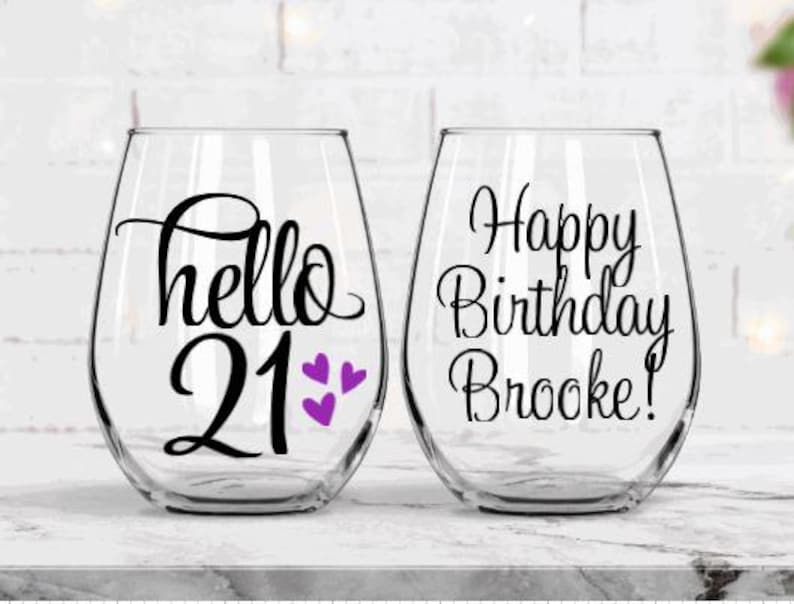 May include: Two clear stemless wine glasses. One glass has the words "hello 21" with purple heart accents. The other glass says "Happy Birthday Brooke!" in black script. The glasses are on a white marble surface.