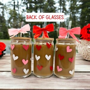May include: Three glass cups with brown liquid and pink and white hearts printed on the outside. Each cup has a bamboo lid and a clear drinking straw with a pink, red, or white bow at the top. The cups are on a wooden table. The text "Back of Glasses" is at the top of the image.