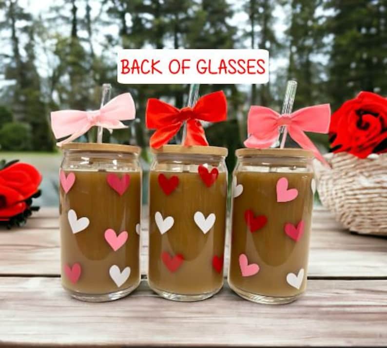 May include: Three glass cups with brown liquid and pink and white hearts printed on the outside. Each cup has a bamboo lid and a clear drinking straw with a pink, red, or white bow at the top. The cups are on a wooden table. The text "Back of Glasses" is at the top of the image.
