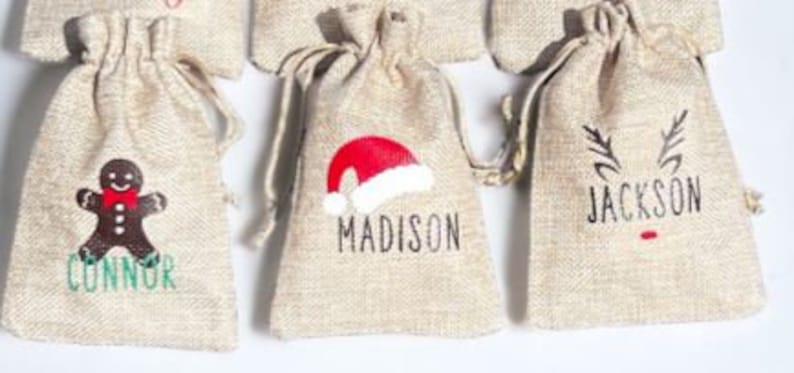 May include: Three small burlap gift bags with personalized names and Christmas-themed designs. The bags are decorated with a gingerbread man, a Santa hat, and reindeer antlers. The names on the bags are Connor, Madison, and Jackson.